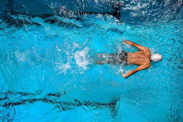Top view of a swimmer wearing a cap, performing a front crawl stroke in a clear blue swimming pool.